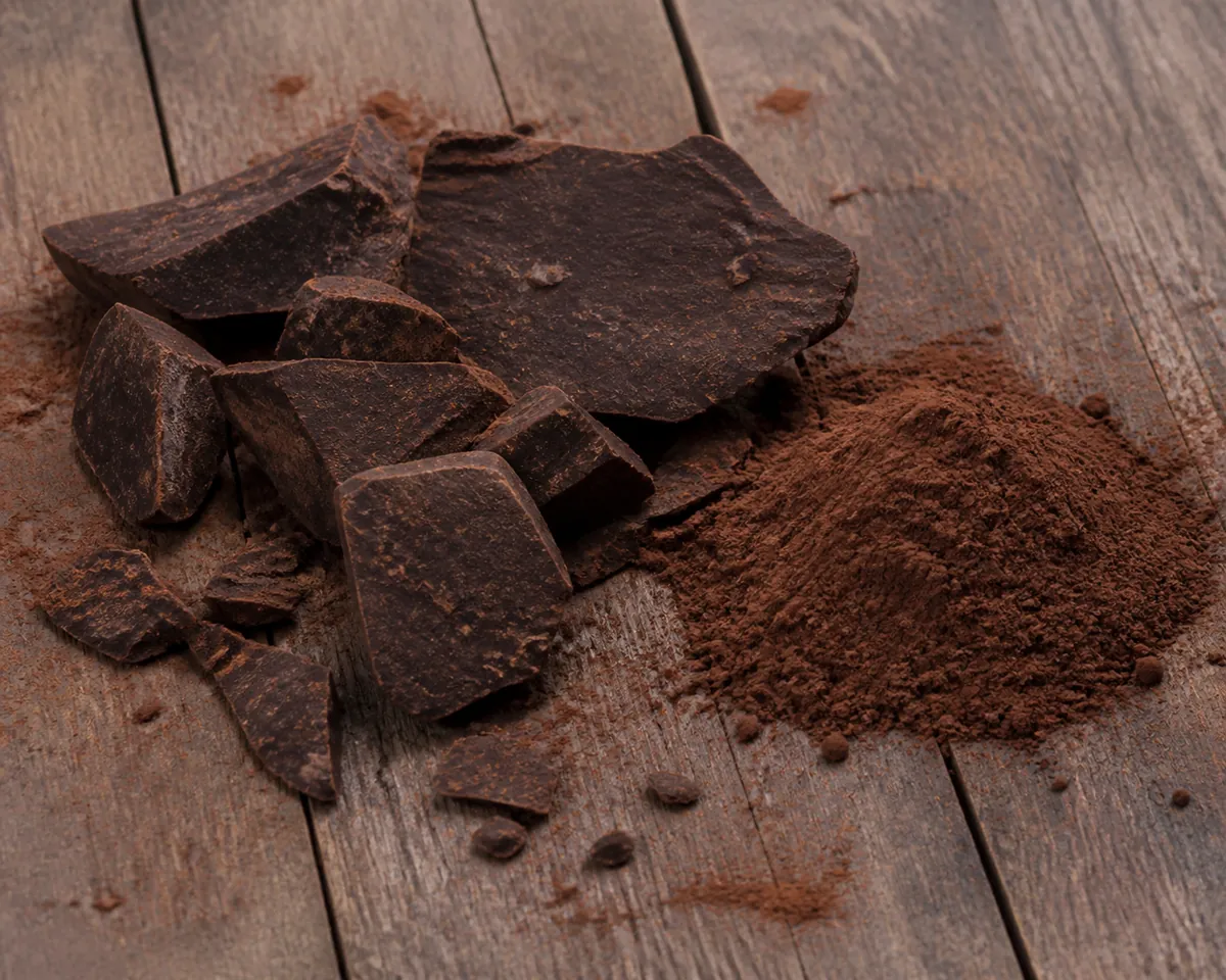 A dark compressed cake of pressed cocoa solids broken into chunks beside a pile of milled cocoa powder on weathered wood
