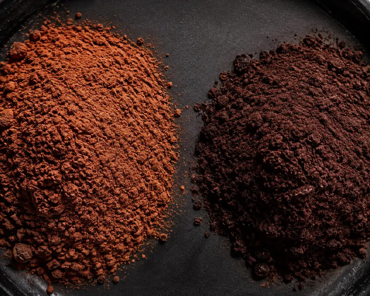 A heap of light reddish-brown natural cocoa powder beside a heap of much darker Dutch-process cocoa on a dark dish