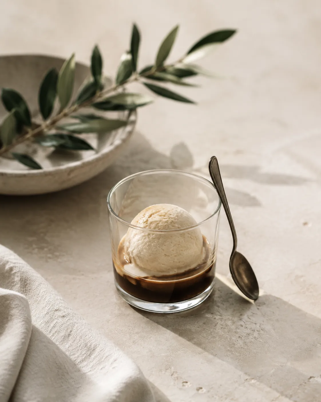 A small clear glass holding a single pale scoop half covered by dark coffee, a short spoon resting against the rim