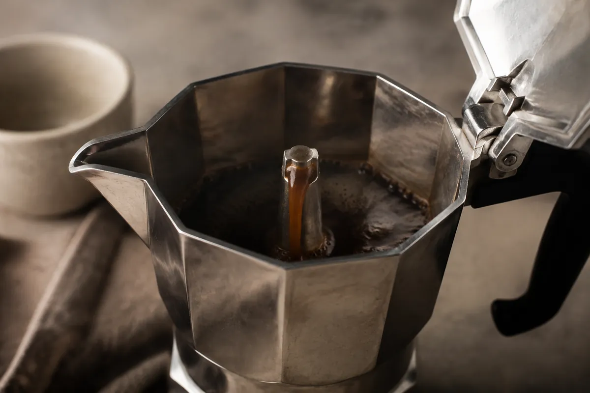 Fresh moka pot coffee emerging in a dark stream from the central chimney into the open upper chamber
