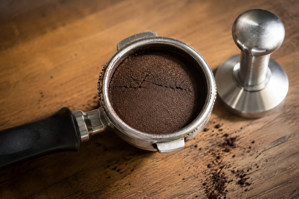 A used espresso puck in a portafilter basket, its surface smooth and domed with a fine crack across it, a tamper resting beside