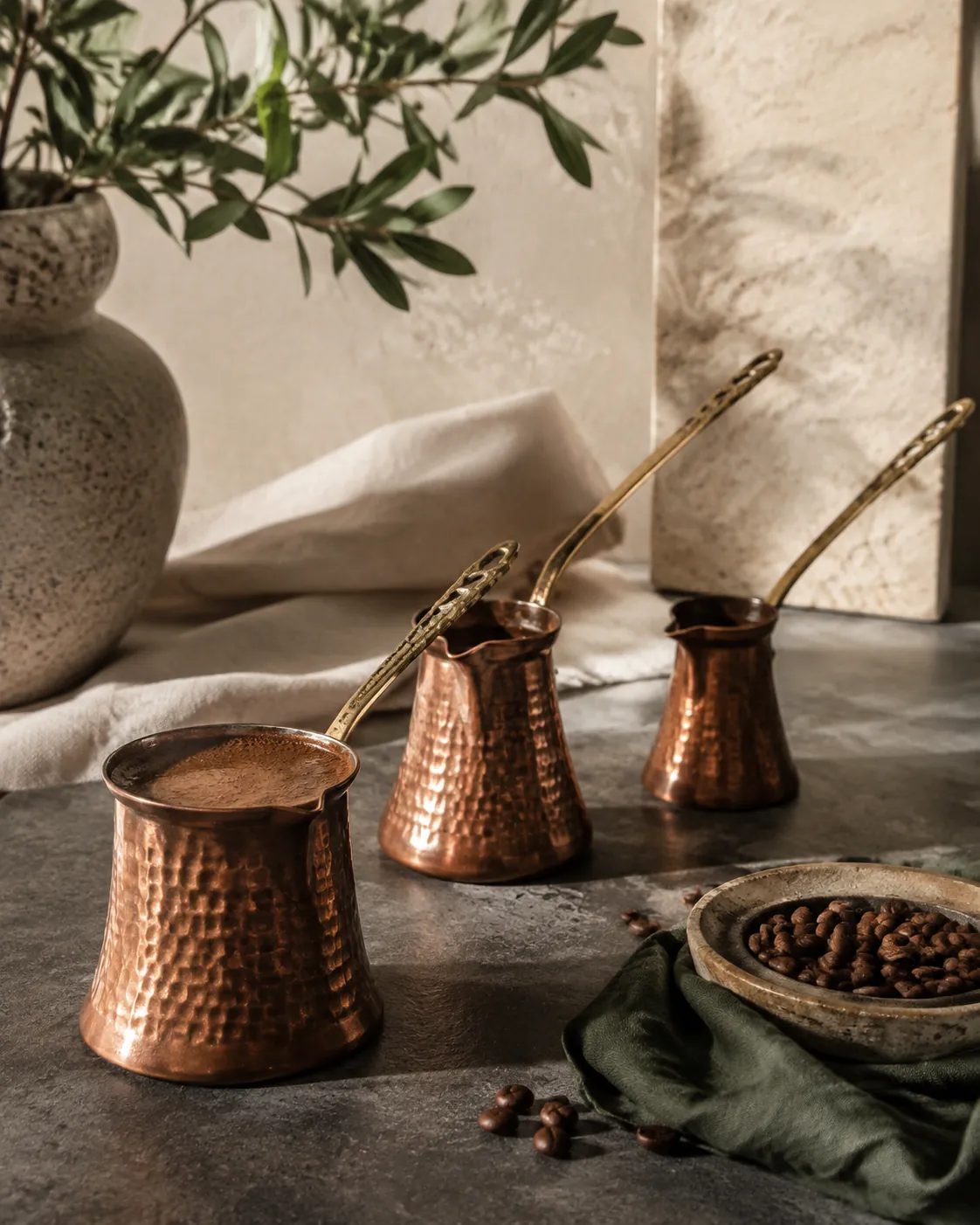 Three long-handled copper cezve pots of different sizes on a dark surface, one with coffee rising in it