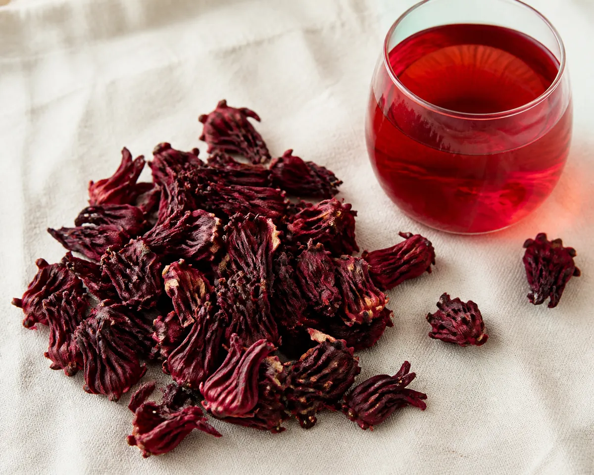 Dried deep red hibiscus calyces heaped on pale linen beside a glass of ruby red infusion
