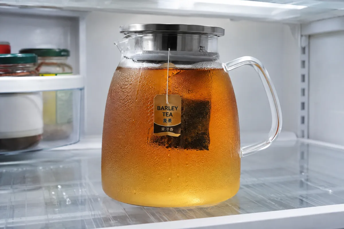 A large glass jug of clear amber barley tea in a fridge with a teabag suspended in it and condensation on the glass
