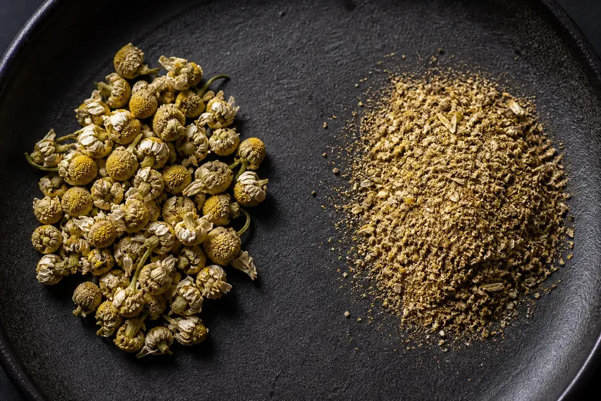 Two piles on a dark ceramic dish: intact dried chamomile flower heads with visible petals on the left, fine brownish chamomile fragments and dust on the right