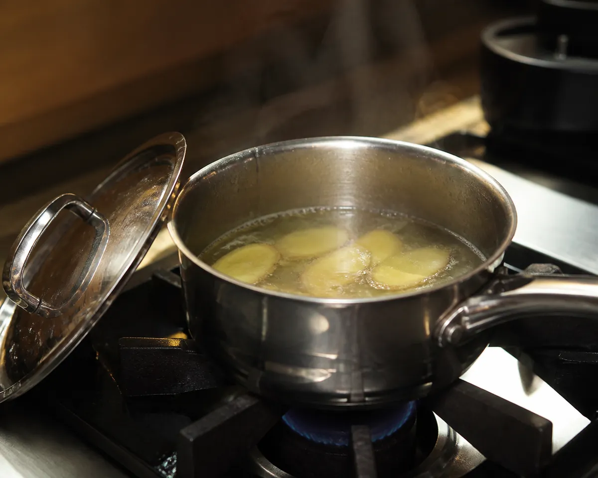 A small stainless steel pan on a stove holding water and thin ginger coins at a gentle simmer, with the lid resting tilted against the pan