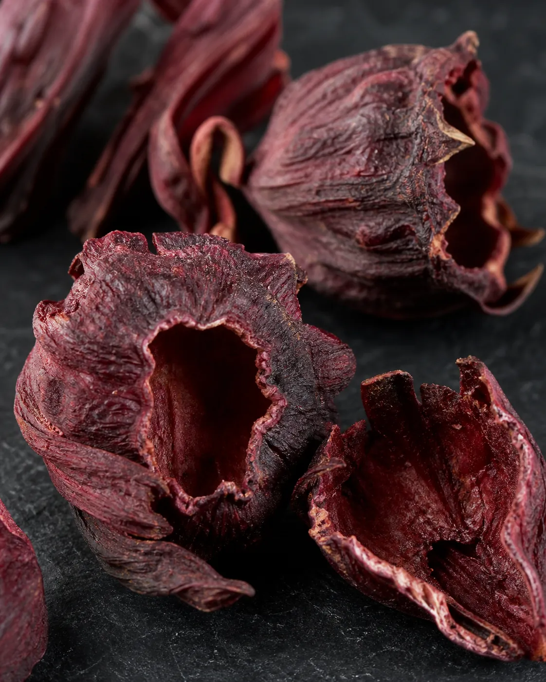 Close macro of dried hibiscus calyces on dark slate showing the cup shape and deep crimson wrinkled surface, one split open