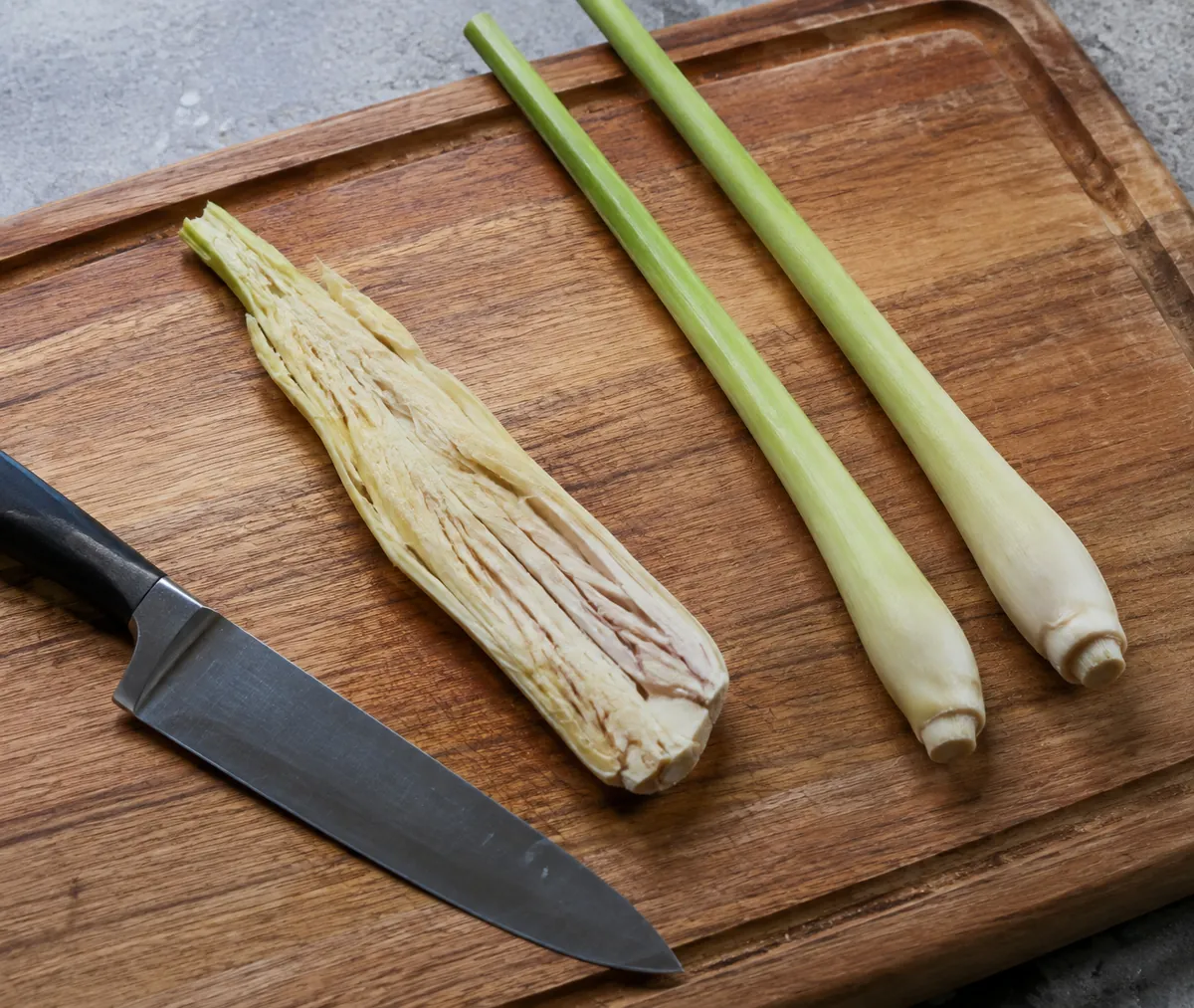 A trimmed pale lemongrass stalk crushed flat along its length with the fibrous layers splayed open, beside two untrimmed green stalks and a knife