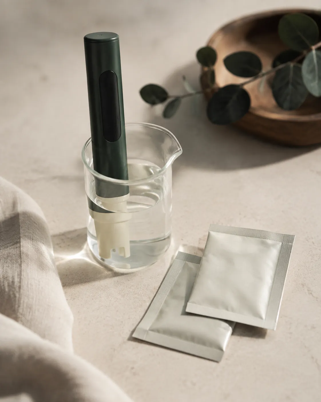 A slim digital pH meter standing in a small glass beaker of clear water, two calibration sachets beside it on a pale surface
