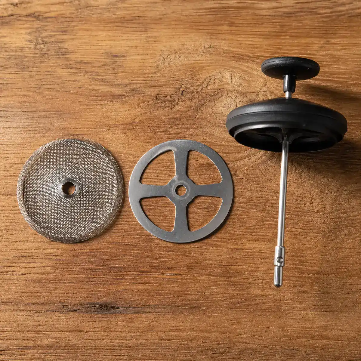 A disassembled French press plunger showing the metal mesh screen and cross plate laid flat on a wooden board