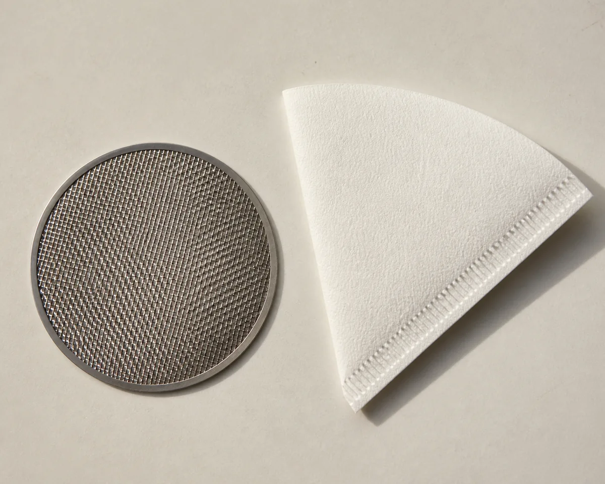 A metal mesh filter disc and a folded paper filter cone laid side by side on a pale surface