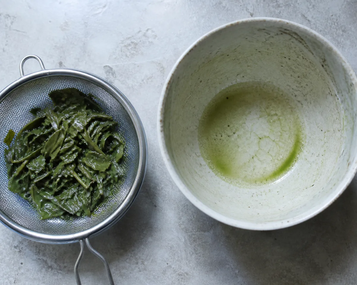Spent wet green tea leaves in a strainer beside an empty matcha bowl with only a green film left