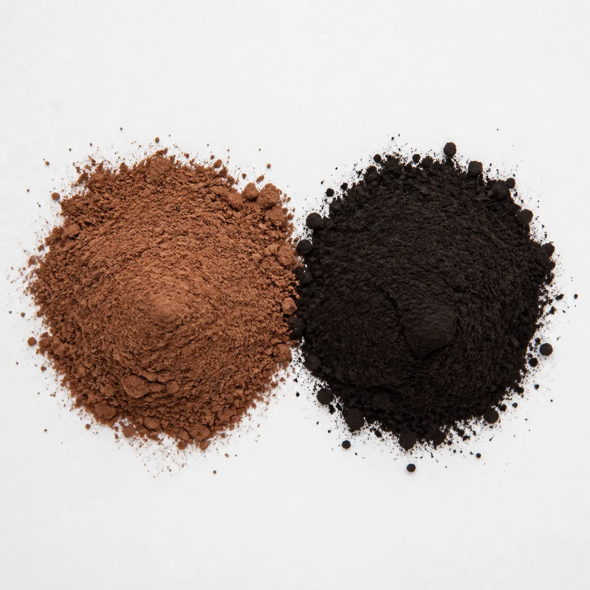 Two small mounds of cocoa powder side by side on white paper, one pale reddish-brown and one dark near-black