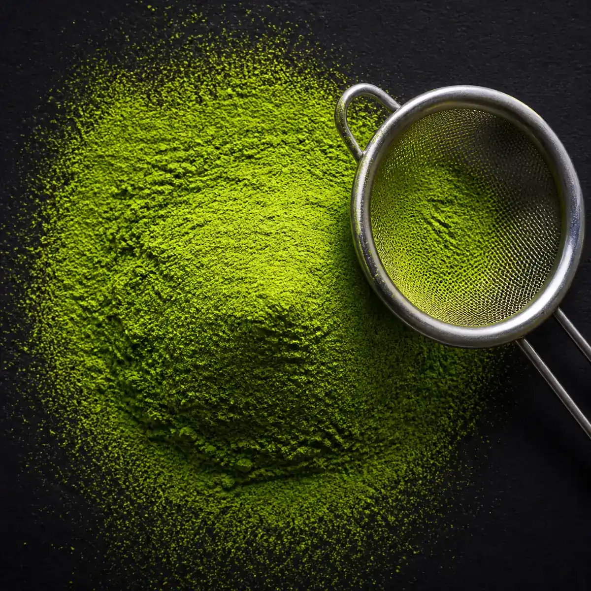 A mound of vivid green matcha powder sifted onto dark paper, with a fine sieve resting alongside