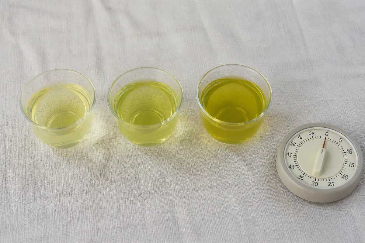 Three small glass cups of green tea in a row showing progressively darker yellow-green liquor, a simple analogue timer lying beside them