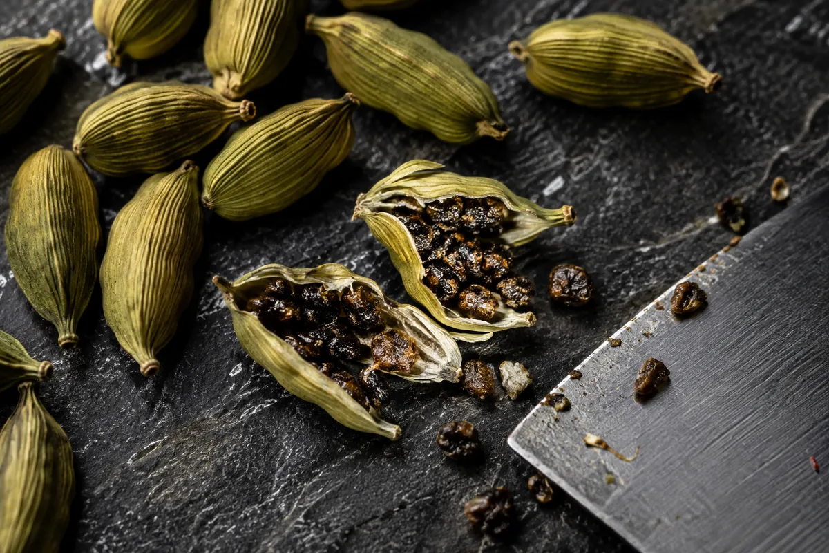 Green cardamom pods on dark slate with two crushed open beside a knife blade, showing the sticky black seeds spilling out