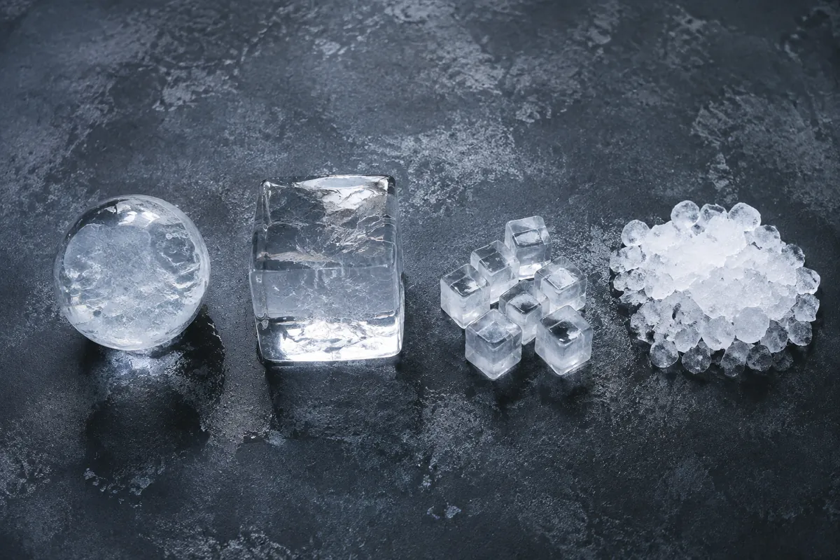 Four forms of ice in a row on a chilled dark surface: a large sphere, a large cube, small standard cubes, and a heap of crushed ice