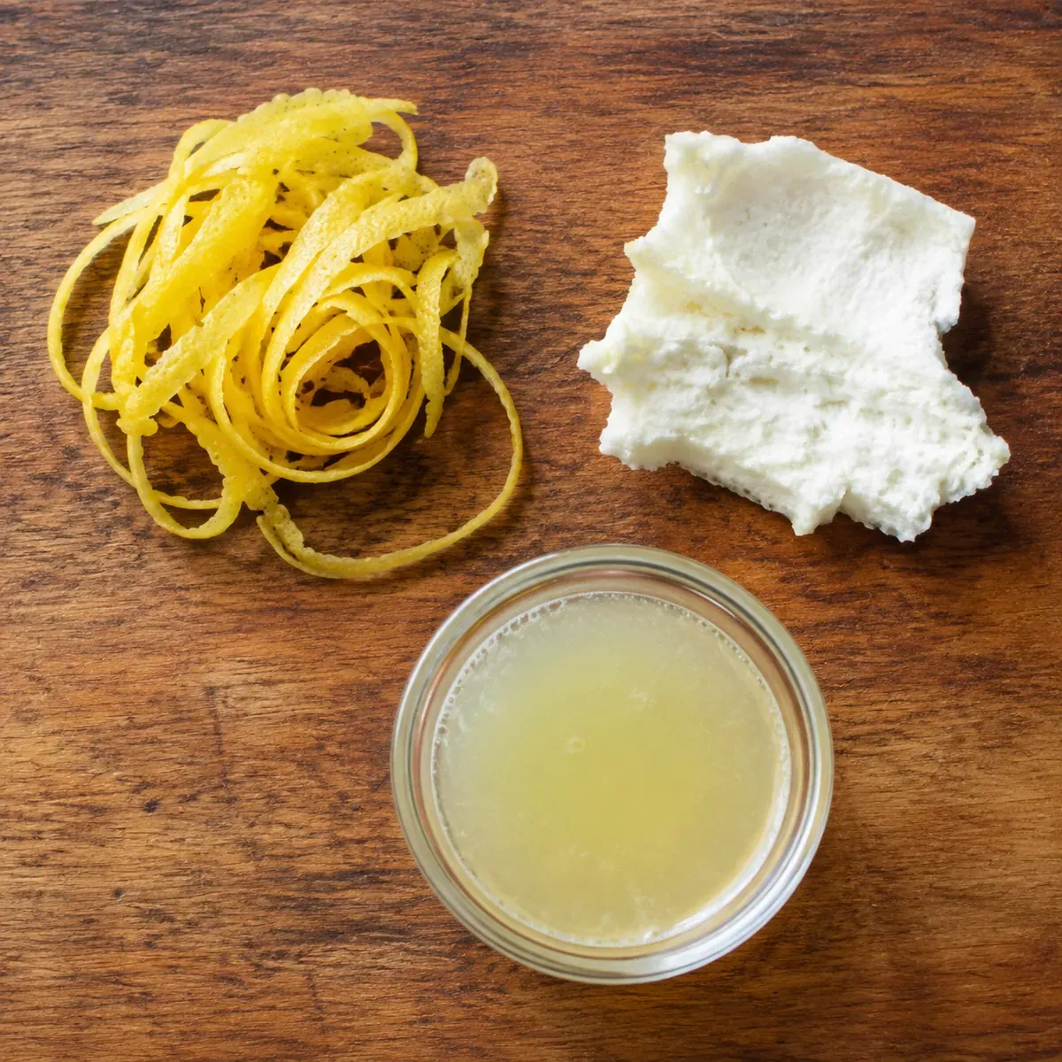 A lemon on a board with a curl of yellow zest, a piece of white pith and a small glass of juice arranged apart from each other
