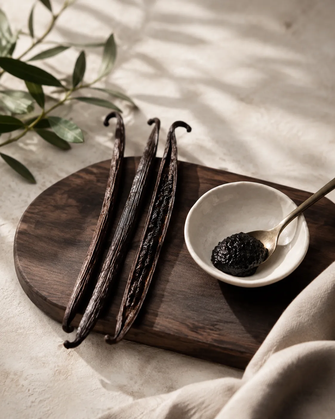 Three whole dark vanilla pods with one split lengthwise to show the seeds, arranged beside a small dish of vanilla paste on a dark wooden board