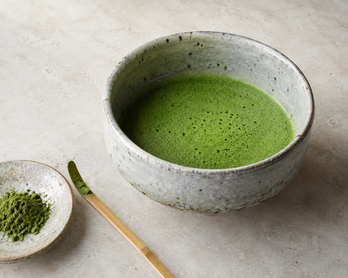 A bowl of whisked matcha beside a small dish of matcha powder and a scale