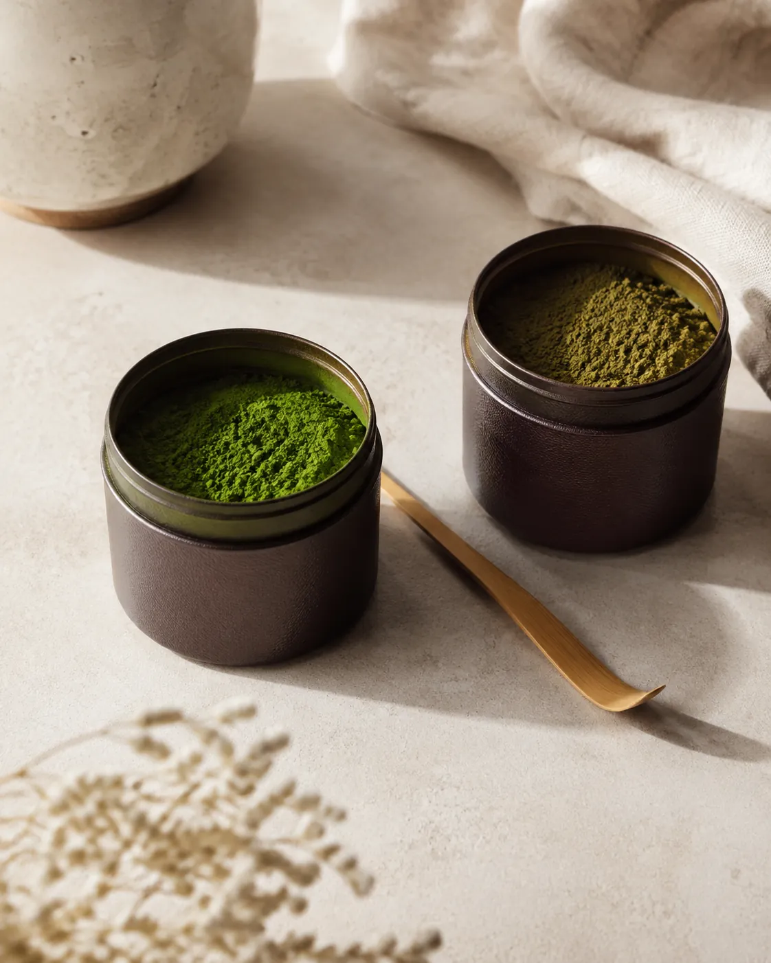 Two small opaque matcha tins open on a bench, the powder in each a slightly different green, a bamboo scoop between them