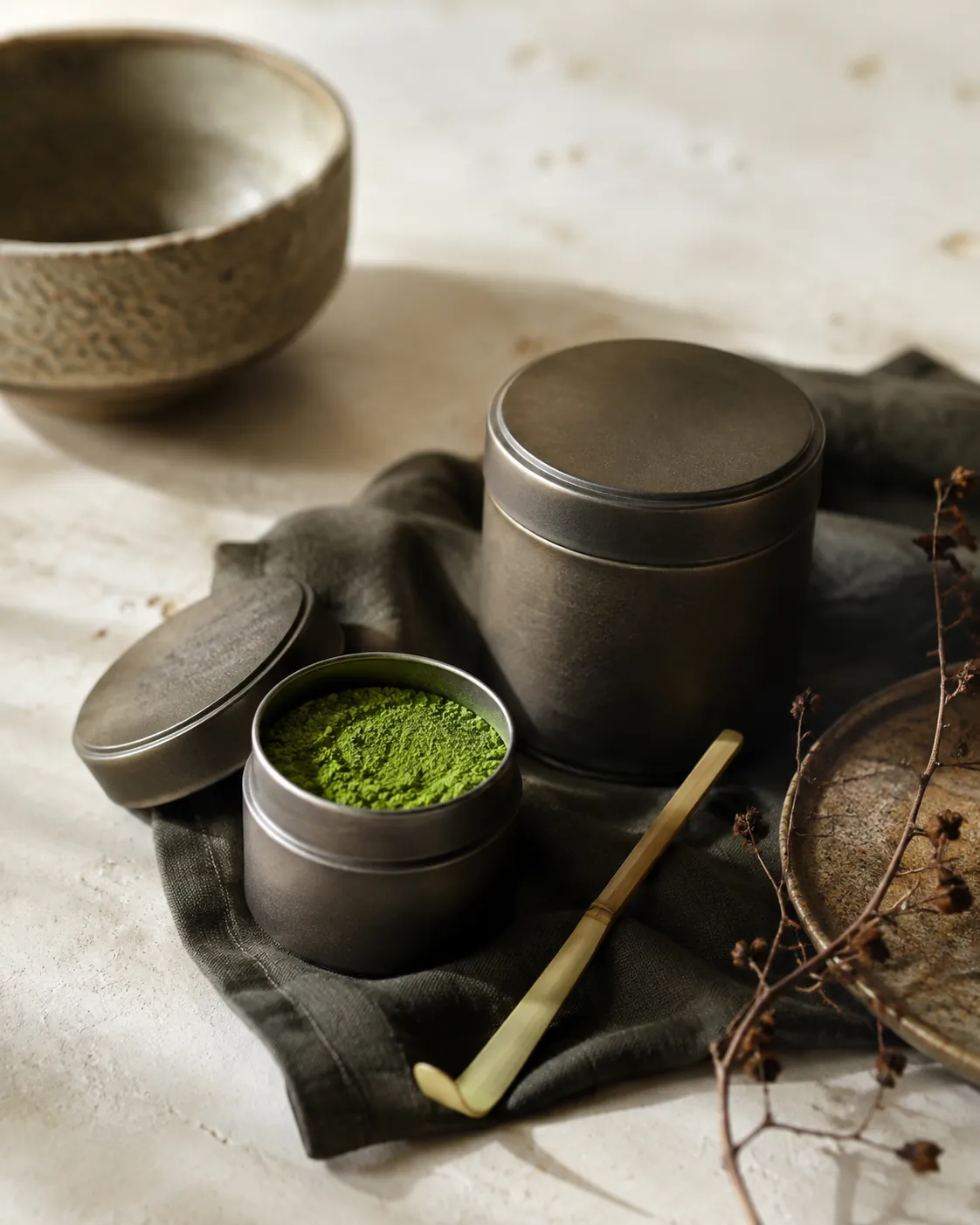 A small tin of vivid green matcha open beside a larger sealed tin, a bamboo scoop resting between them on dark linen