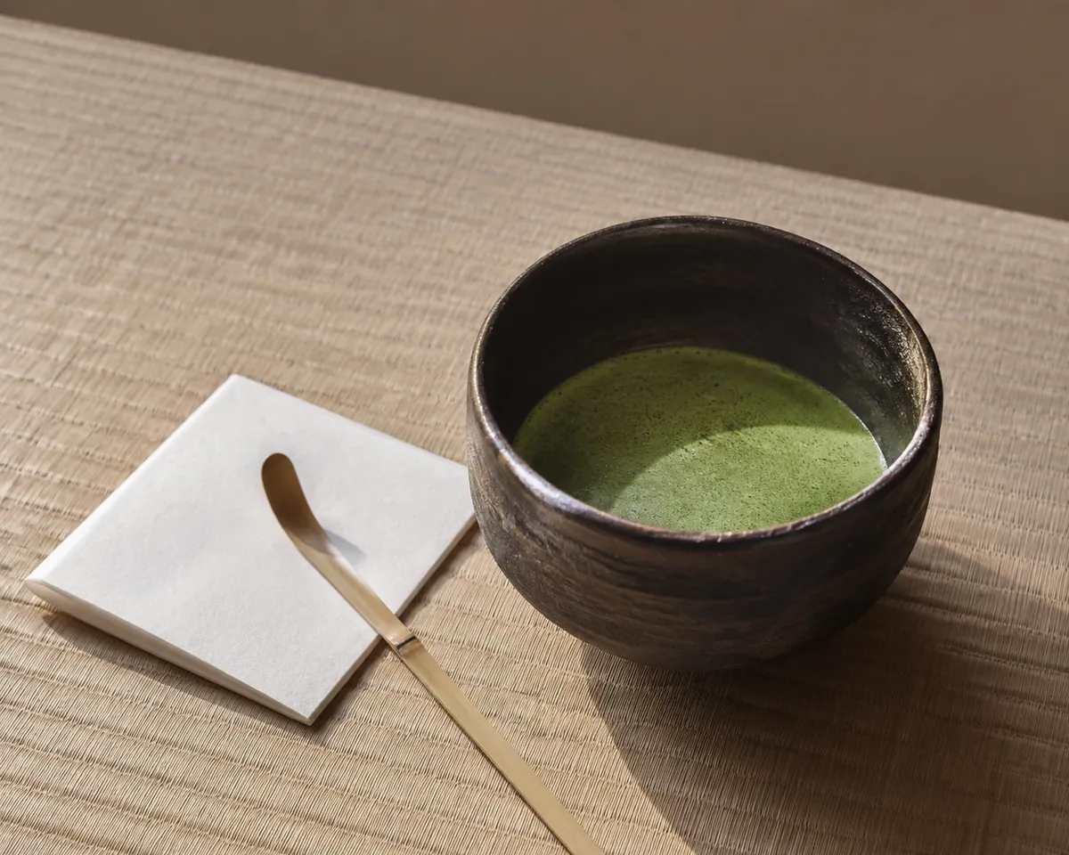 A large dark tea bowl of thick matcha resting on tatami with a folded paper square beside it