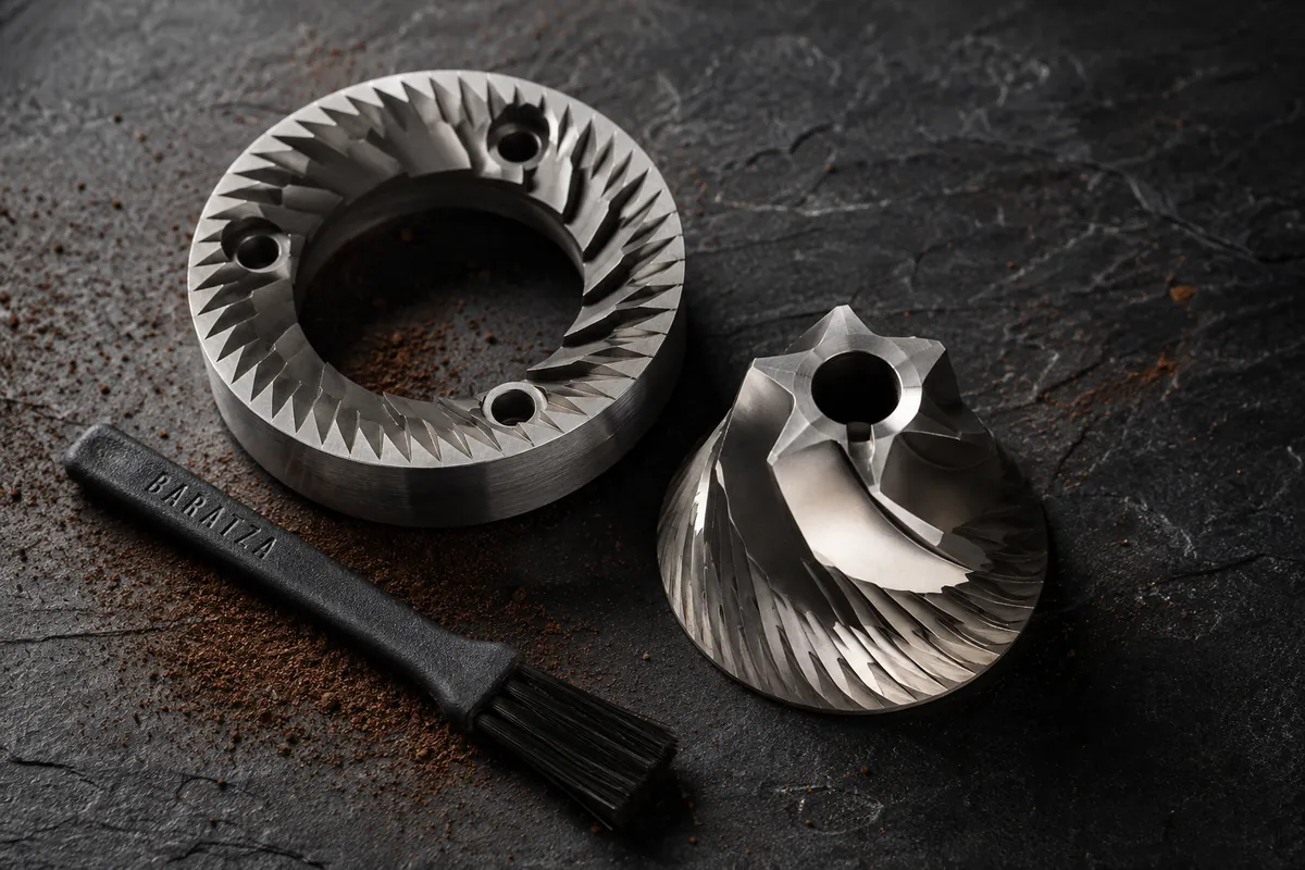 A conical steel grinder burr and its outer ring resting separated on dark slate with a brush beside them and a dusting of fine coffee