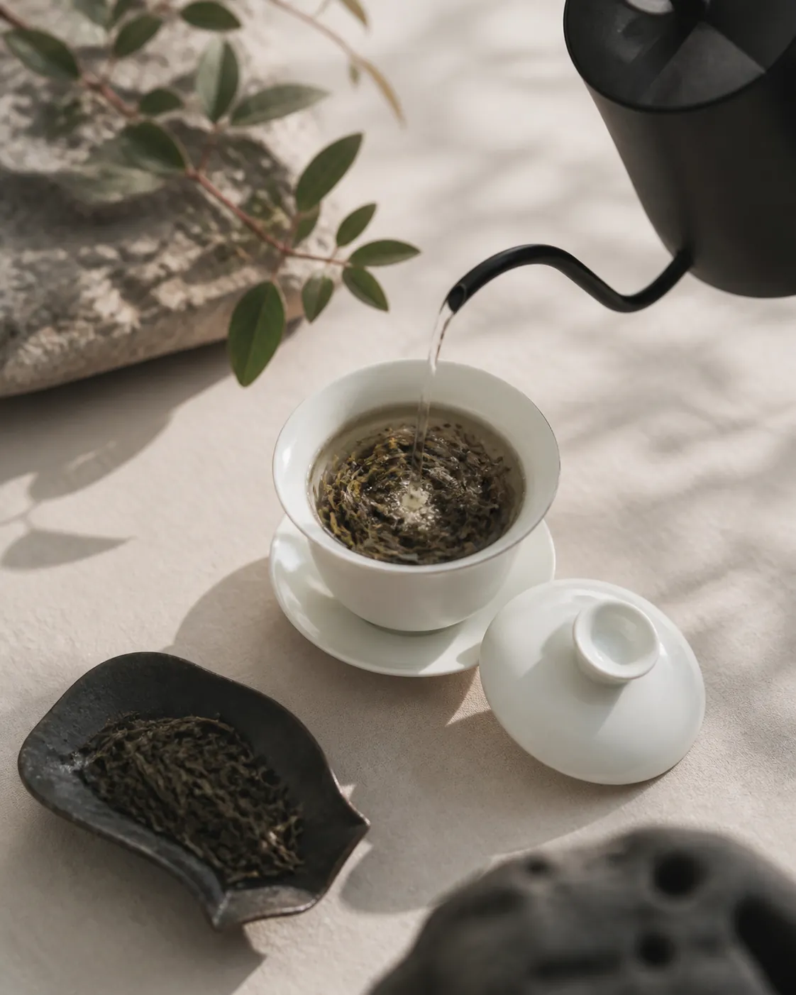 A gooseneck kettle filling a small porcelain gaiwan from a height, leaf turning in the water