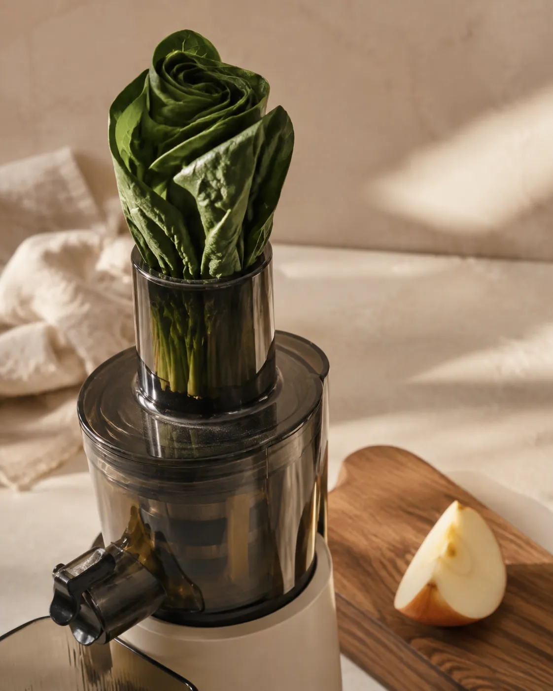 A bundle of rolled spinach leaves being fed into a juicer chute beside a wedge of apple on a wooden board