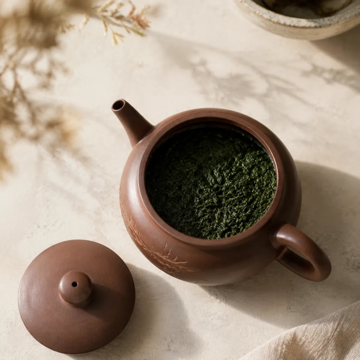 An open teapot showing a dense bed of fine dark green wet tea leaf filling the base