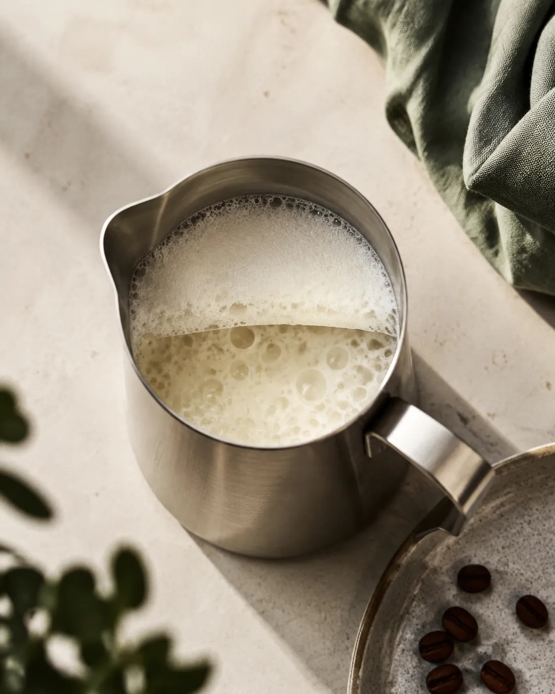 A steel milk jug of milk covered in large loose bubbles with a dry stiff foam lid sitting apart from the liquid beneath