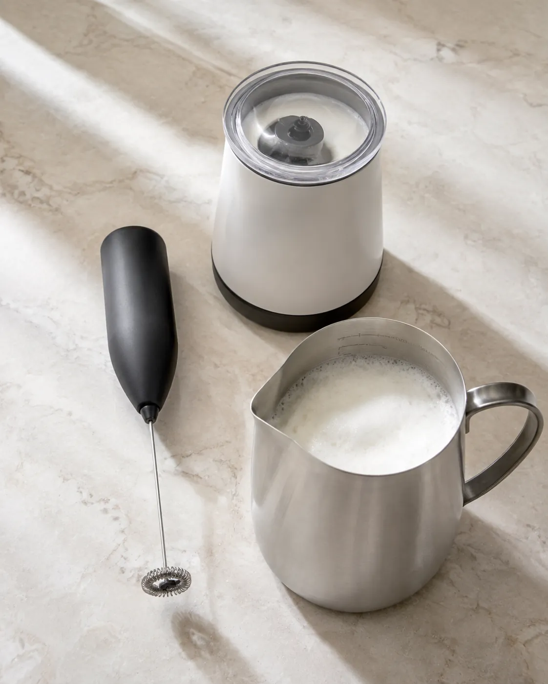 A handheld milk frother beside an automatic jug frother and a steel milk jug