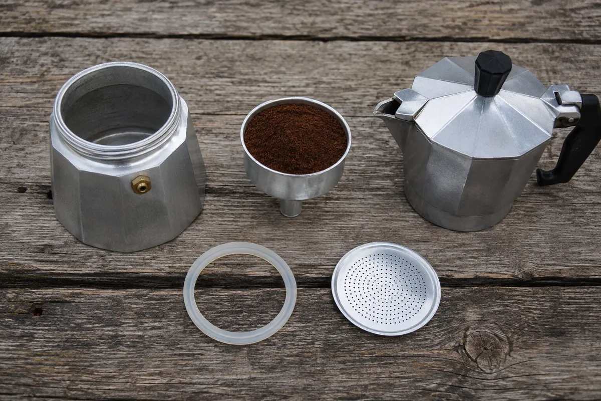 An aluminium stovetop pot disassembled into base, funnel basket filled with ground coffee, and top chamber, with gasket and filter plate beside them