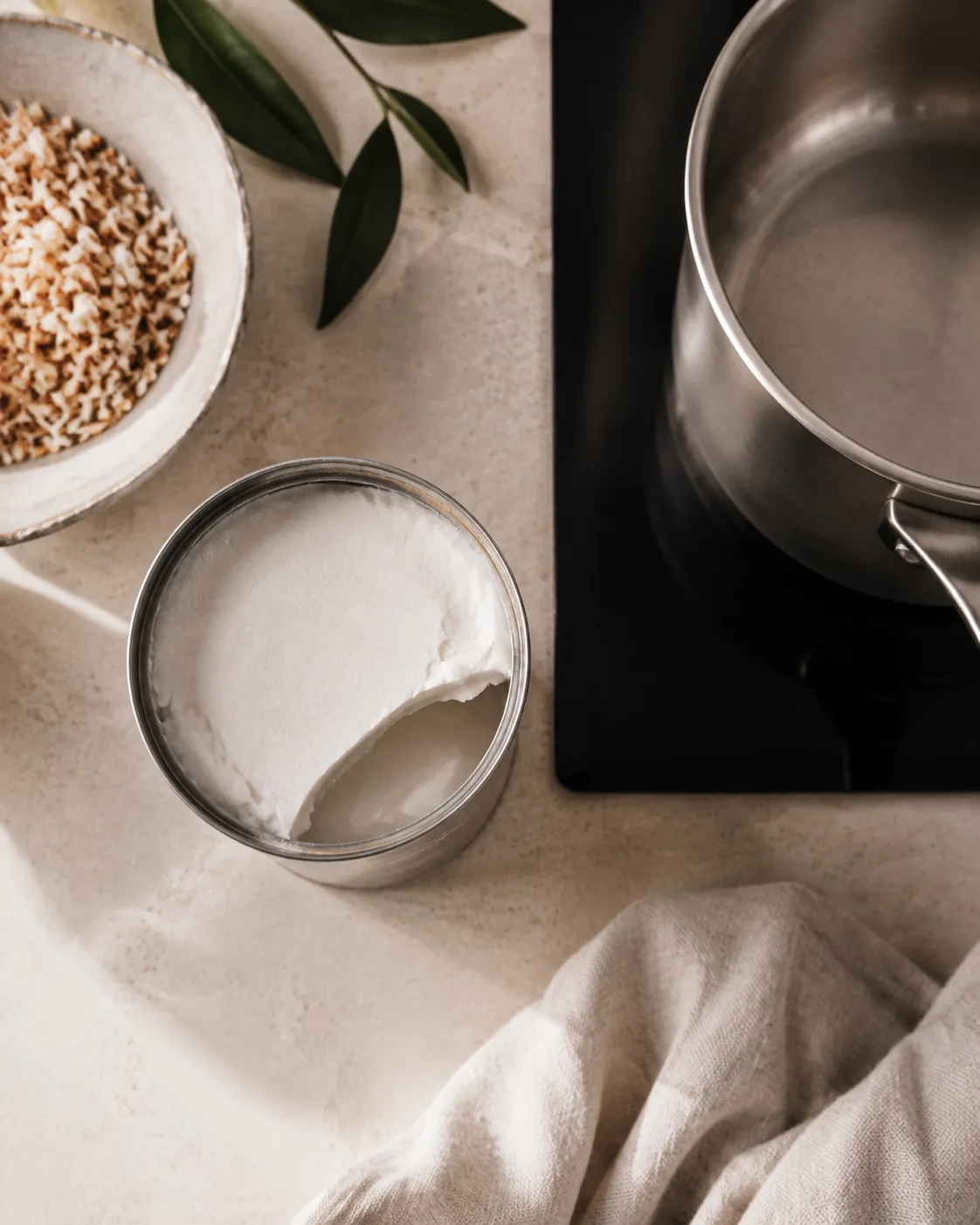 An opened plain unlabelled tin of coconut milk seen from above, thick white cream sitting on thinner liquid below, beside a small saucepan