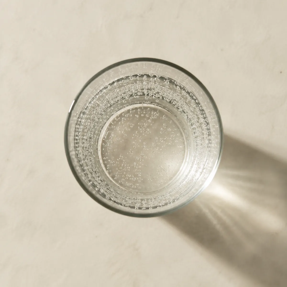 A plain glass of clear carbonated water seen from directly above on a bare white surface, fine bubbles clinging inside the glass