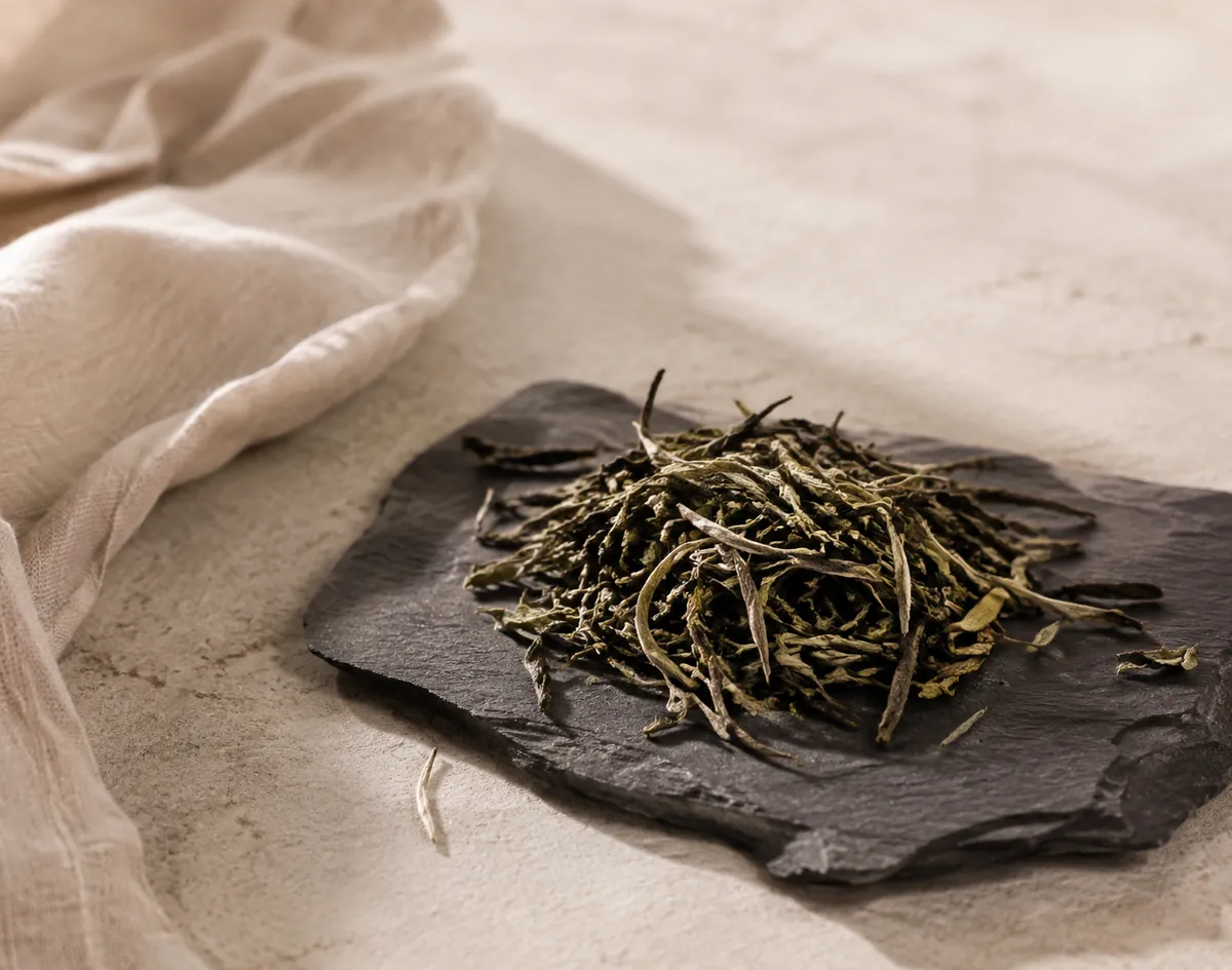 A small heap of dry Darjeeling first flush leaf on dark slate, greenish grey and wiry with pale silver tips scattered through it