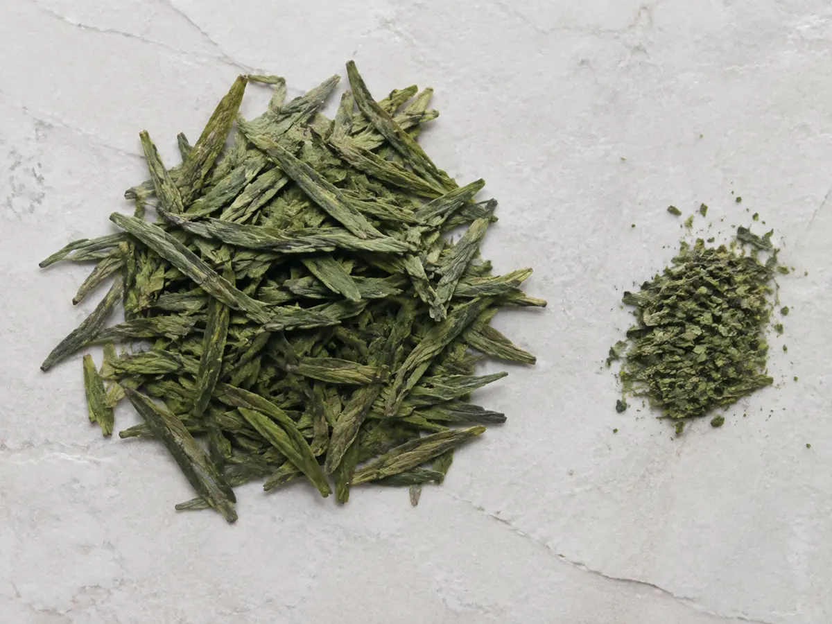 Flat jade-green Dragon Well tea leaves spread beside a smaller pile of broken leaf and dust
