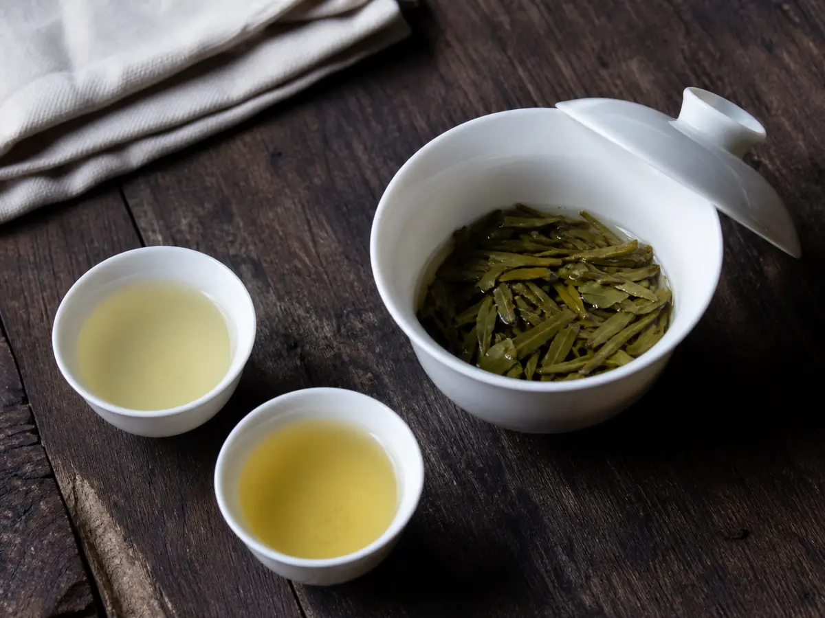 Open porcelain gaiwan with softened Dragon Well leaves beside two pale yellow-green infusions