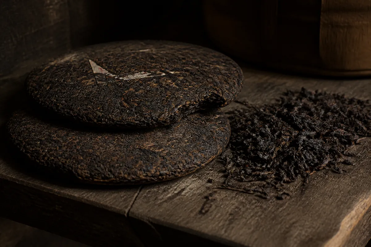 Two compressed discs of aged dark oolong stacked on a weathered wooden shelf beside a heap of dark glossy roasted leaf, in warm dim storage light