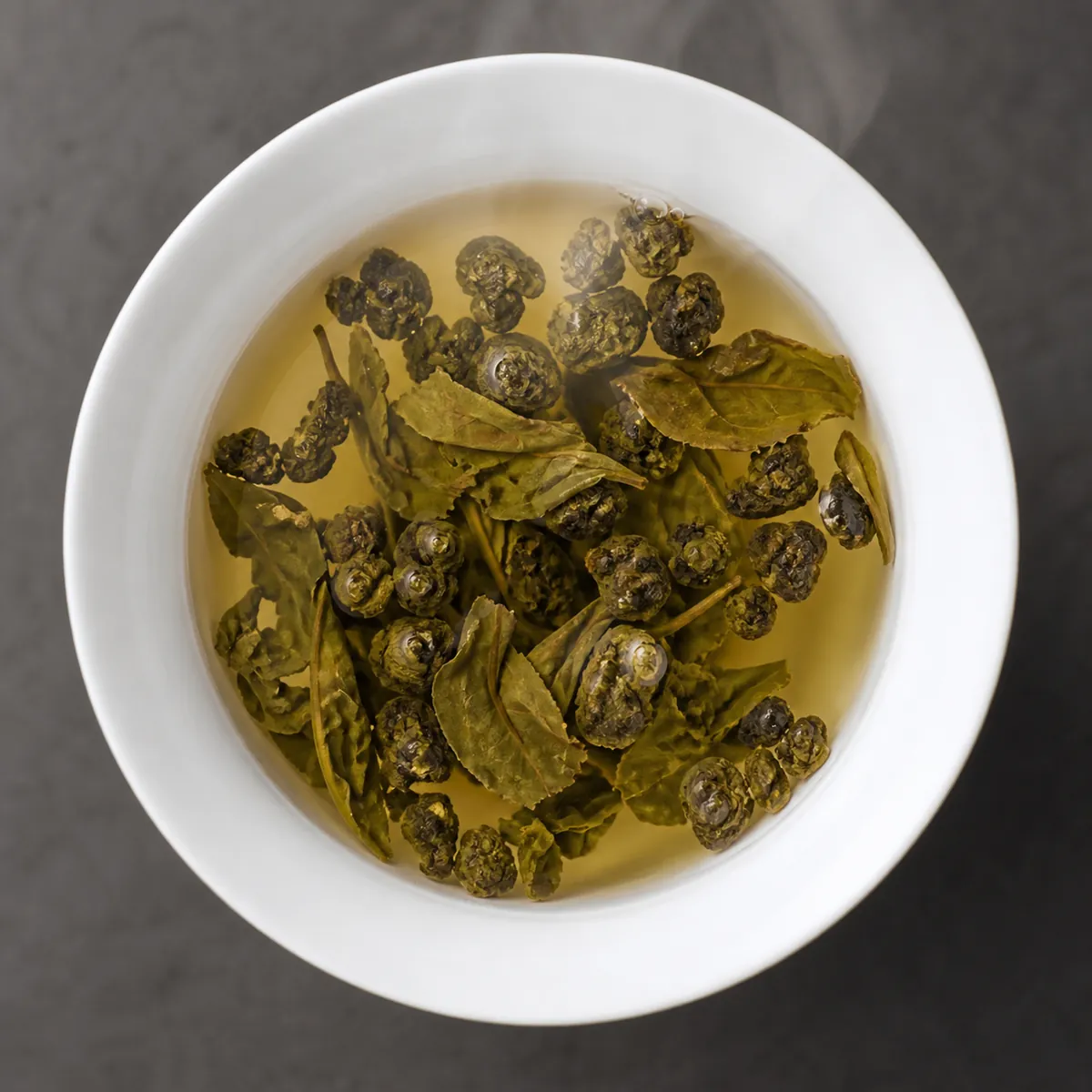 A porcelain gaiwan seen from above holding rolled oolong pellets mid-way through opening, some still tight balls and some unfurled into whole leaves